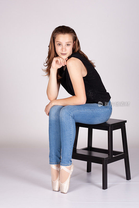 En Pointe坐在凳子腿上，腼腆的表情看着镜头，手到下巴，耸肩，漂亮的十几岁的女孩，看起来很好奇，有马基雅维利主义的倾向